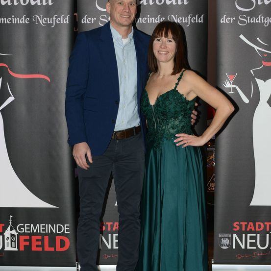 Ein Mann und eine Frau stehen lächelnd nebeneinander in formeller Kleidung. Hinter ihnen befinden sich zwei Banner mit den Texten 'Gemeinde Neufeld' und 'Stadt Neufeld'.