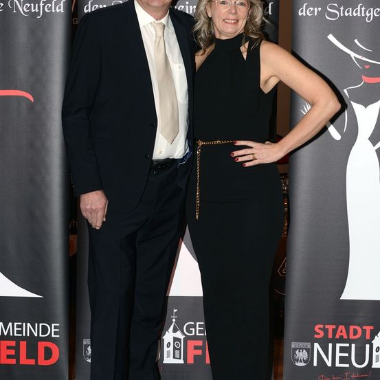 Ein Mann und eine Frau stehen zusammen und sind in formeller Kleidung gekleidet. Der Mann trägt einen schwarzen Anzug und eine beige Krawatte, während die Frau ein schwarzes Kleid mit einem goldenen Gürtel trägt. Sie lächeln beide und scheinen für ein Foto zu posieren. Hinter ihnen befinden sich zwei Banner, von denen eines das Gesicht einer Frau zeigt.