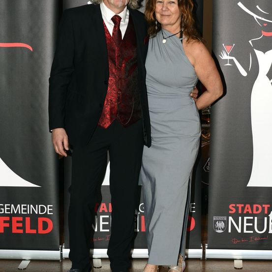 Ein Mann in einem schwarzen Anzug und einer roten Weste steht neben einer Frau in einem grauen Kleid, beide lächeln. Sie stehen vor einem Banner mit einem Stadtlogo.