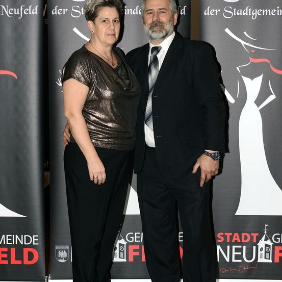 Ein Mann und eine Frau stehen eng beieinander auf einem roten Teppich bei einer Veranstaltung und lächeln für ein Foto. Hinter ihnen sind Banner mit dem Text Stadt Neufeld und GEI F.
