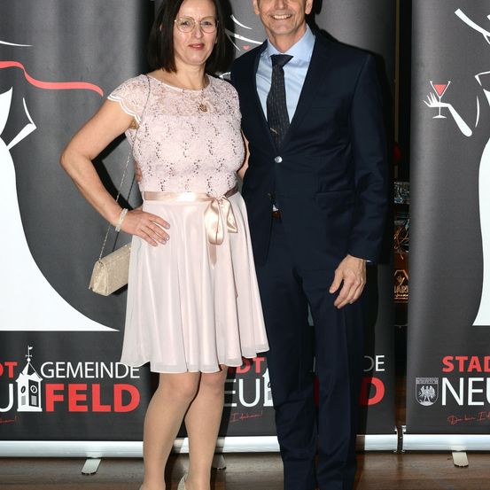 Eine Frau und ein Mann, schick gekleidet, stehen bei einer Veranstaltung zusammen. Die Frau trägt ein rosa Kleid und der Mann einen dunklen Anzug. Sie stehen vor einem Banner mit Text und einer Illustration.