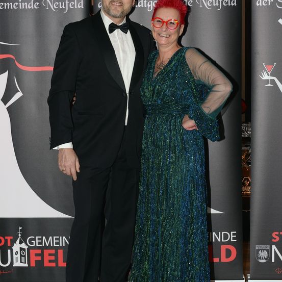 Ein Mann und eine Frau stehen gemeinsam auf einem roten Teppich und lächeln für die Kamera. Der Mann trägt einen Frack und die Frau ein grünes Paillettenkleid. Sie sind von Veranstaltungsbannern umgeben.