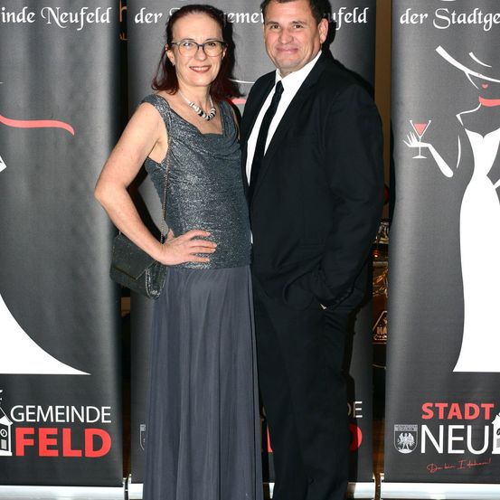 Ein Mann und eine Frau in formeller Kleidung stehen lächelnd nebeneinander vor einem Hintergrund.