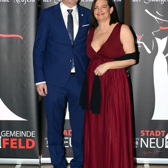 Ein Mann und eine Frau stehen nebeneinander und lächeln vor einem Hintergrund mit Logos und Text.