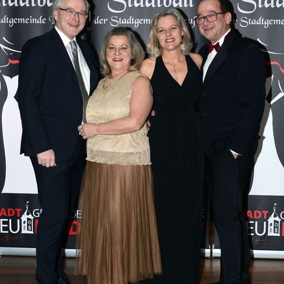 Vier Personen, zwei Männer und zwei Frauen, stehen gemeinsam auf einer Bühne. Sie sind feierlich gekleidet, wobei die Frauen elegante Kleider tragen. Im Hintergrund befindet sich ein Banner mit Text in deutscher Sprache, was auf eine Veranstaltung oder Preisverleihung hindeutet.