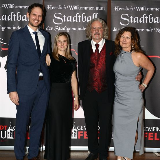 Vier Personen, zwei Männer und zwei Frauen, stehen gemeinsam vor einer Kulisse. Sie sind feierlich gekleidet. Der Mann links trägt einen blauen Anzug, und die Frau neben ihm trägt ein schwarzes Kleid. Der Mann in der Mitte trägt ein rotes Weste und eine Brille. Die Frau rechts trägt ein graues Kleid und hat ihren Arm um die Schulter des Mannes gelegt.