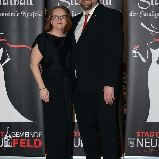 Ein Mann und eine Frau in formeller Kleidung stehen gemeinsam vor einem Banner der Gemeinde Neufeld.