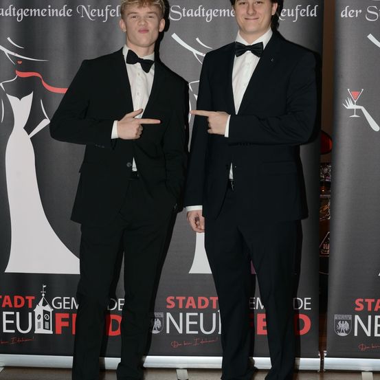 Zwei junge Männer in formellen Anzügen stehen nebeneinander, einer zeigt und der andere lächelt, vor einem Banner.