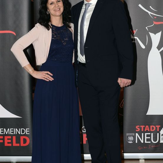 Ein Mann und eine Frau stehen Seite an Seite in formeller Kleidung und lächeln für ein Foto. Die Frau trägt ein marineblaues Kleid und eine beige Jacke, während der Mann einen schwarzen Anzug mit Krawatte trägt. Sie stehen vor einem Hintergrund mit dem Text 'Stadt Neu'.