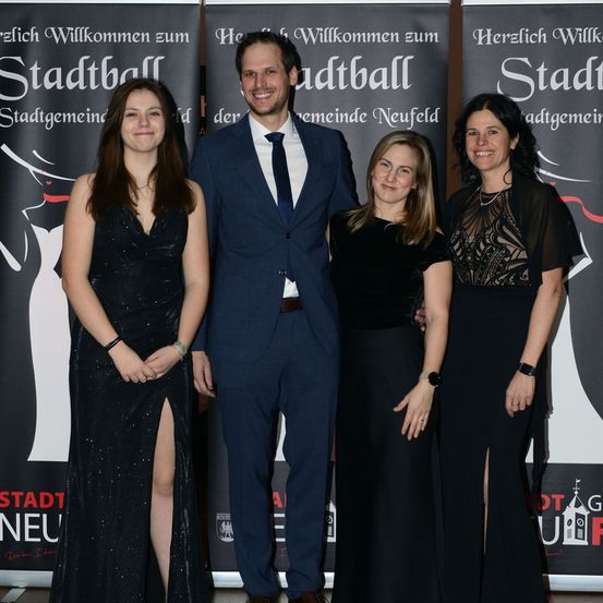 Vier Personen, zwei Frauen und zwei Männer, stehen zusammen vor einem schwarzen Hintergrund mit rotem Text. Sie lächeln und scheinen für ein Foto zu posieren.