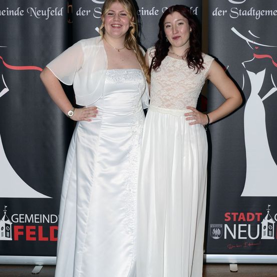 Zwei Frauen in weißen Kleidern stehen lächelnd nebeneinander vor einem Werbebanner. Eine trägt ein Spitzenoberteil, die andere ein ärmelloses Kleid. Beide haben Accessoires.