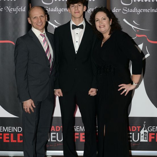 Drei Menschen stehen eng beieinander vor einem Banner und lächeln für ein Foto. Der Mann links trägt einen Anzug mit einer gestreiften Krawatte. Der Mann in der Mitte, der einen Frack mit einer Fliege trägt, steht zwischen zwei Frauen. Die Frau rechts trägt ein schwarzes Kleid mit den Händen auf den Hüften.