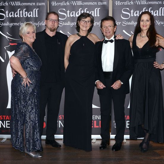 Eine Gruppe von Menschen, die feierlich gekleidet sind, steht gemeinsam vor einem schwarzen Banner mit dem Wort 'Stad' darauf. Sie lächeln und scheinen für ein Foto zu posieren.