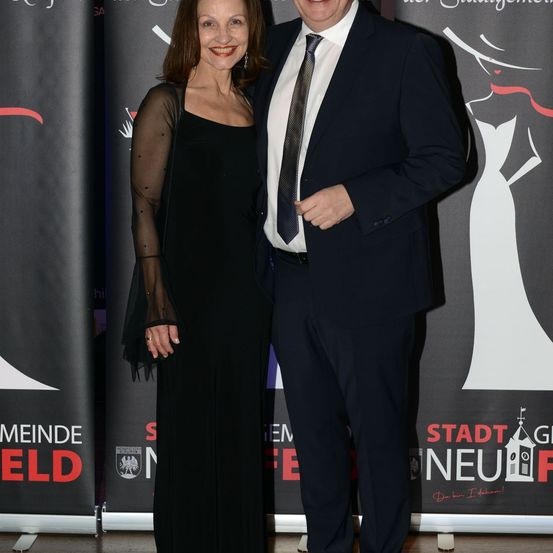 Ein Mann und eine Frau stehen nebeneinander bei einer Veranstaltung. Der Mann trägt einen Anzug und eine Krawatte, und die Frau trägt ein schwarzes Kleid. Sie beide lächeln und schauen in die Kamera. Hinter ihnen ist ein Hintergrund mit dem Text 'Stadt Neuf'.