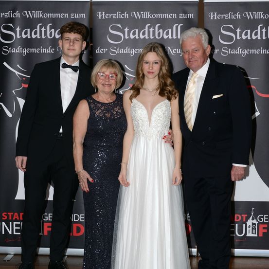 Vier Personen stehen vor einem Banner, zwei Männer und zwei Frauen. Die Frauen tragen Kleider und die Männer Anzüge. Die Frau im weißen Kleid hat lange Haare.