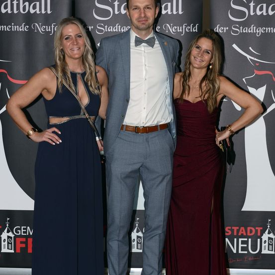 Drei Personen, zwei Frauen und ein Mann, posieren für ein Foto bei einer Veranstaltung. Der Mann trägt eine Fliege und die Frauen elegante Kleider. Sie stehen vor einem Hintergrund mit Veranstaltungsbranding.