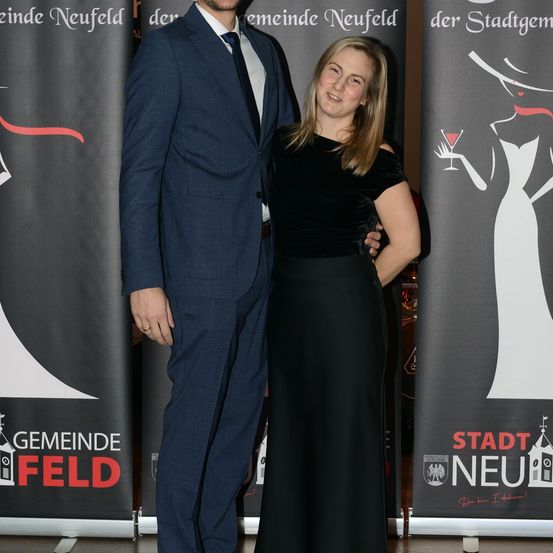 Ein Mann und eine Frau stehen dicht nebeneinander vor einem Banner mit dem Wort Stadt. Der Mann trägt einen blauen Anzug und eine Krawatte. Die Frau trägt ein schwarzes Kleid. Sie lächeln beide und scheinen für ein Foto zu posieren.