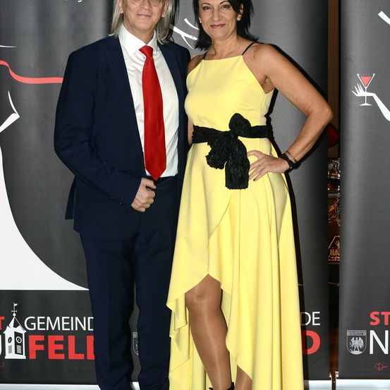 Ein Mann und eine Frau posieren für ein Foto auf dem roten Teppich. Der Mann trägt einen Anzug und eine rote Krawatte, während die Frau ein gelbes Kleid mit einem schwarzen Gürtel trägt.