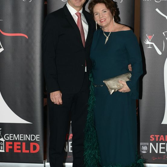 Ein Mann und eine Frau stehen nah beieinander und lächeln. Sie sind bei einer Veranstaltung. Die Frau hält eine Handtasche. Hinter ihnen sind Banner. Ein Banner sagt 'Gemeinde Feld'. Ein anderes sagt 'Stadt Neu'.