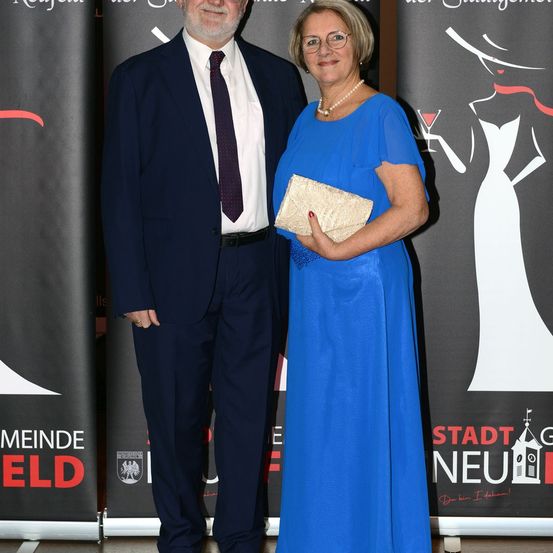 Ein Mann und eine Frau in formeller Kleidung stehen nebeneinander vor einem Hintergrund mit einer stilisierten Frau mit einem Getränk und dem Text Stadt Neu.