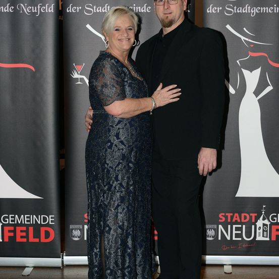 Eine lächelnde Frau in einem blauen Kleid und ein Mann in einem schwarzen Anzug stehen eng nebeneinander vor einem Banner mit deutschem Text.