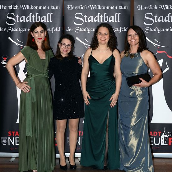 Vier Frauen stehen vor Bannern mit 'Stadtball' darauf. Sie sind alle festlich gekleidet, wahrscheinlich für eine Veranstaltung. Die Frau auf der linken Seite trägt ein grünes Kleid, die Frau neben ihr ein schwarzes Kleid. Die beiden Frauen rechts tragen grüne und graue Kleider.