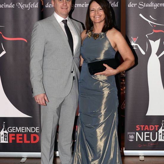 Ein Mann und eine Frau stehen nebeneinander, lächeln und haben ein Banner mit der Aufschrift 'Gemeinde Neufeld' hinter sich. Der Mann trägt einen grauen Anzug und die Frau ein silbernes Kleid.