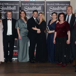 Eine Gruppe von Menschen in formeller Kleidung posiert für ein Foto vor einem Banner, das 'Herzlich Willkommen zum Stadtball' steht.