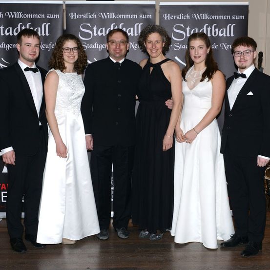 Sechs Personen posieren für ein Foto bei einer Veranstaltung. Sie sind feierlich gekleidet und stehen vor Bannern.