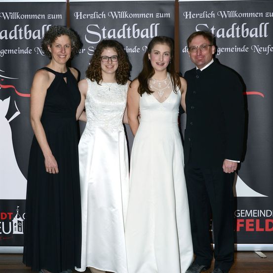 Vier Personen posieren für ein Foto vor einem Banner. Zwei Frauen tragen weiße Kleider und zwei Männer schwarze Anzüge.