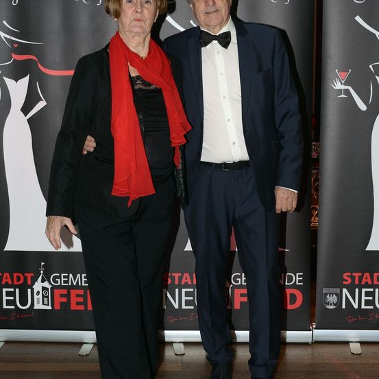 Ein älteres Paar steht vor einem Werbehintergrund mit modernem Architekturthema, beide festlich für eine Veranstaltung gekleidet.