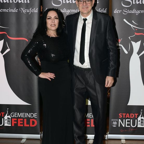 Ein Mann und eine Frau stehen lächelnd zusammen auf einem roten Teppich. Der Mann trägt einen schwarzen Anzug und eine Krawatte, während die Frau ein schwarzes Paillettenkleid trägt. Sie stehen vor Bannern, eines davon zeigt eine Silhouette einer Frau in einem weißen Kleid.