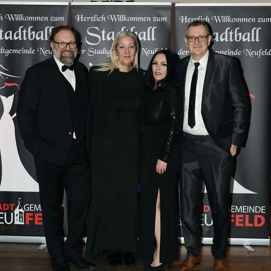 Vier Personen posieren in formeller Kleidung für ein Foto. Sie stehen vor einer Kulisse mit einem Schild, das Stadtball steht.