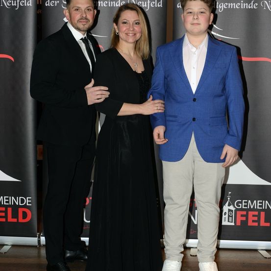 Ein Mann und eine Frau in formeller Kleidung stehen neben einem jungen Jungen in einem blauen Blazer, alle lächeln für die Kamera vor einem Hintergrund mit Gemeinde Neufeld Text.