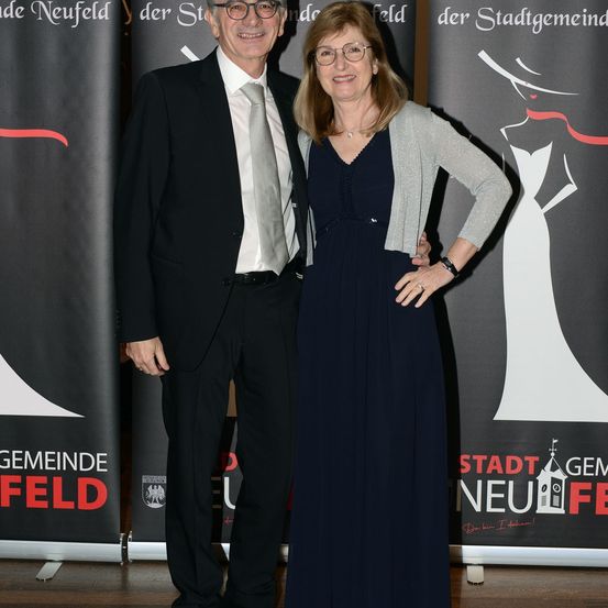 Ein älteres Paar steht lächelnd im Innenraum, mit Bannern im Hintergrund. Der Mann trägt einen schwarzen Anzug und eine Krawatte. Die Frau trägt ein blaues Kleid und eine graue Strickjacke.