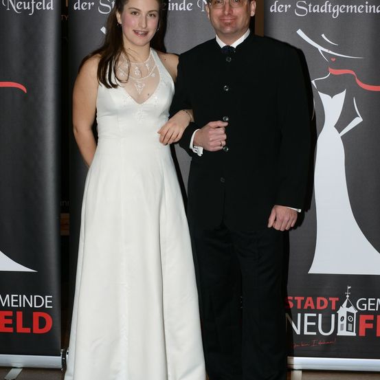 Ein Mann und eine Frau in formeller Kleidung stehen nah beieinander und lächeln für ein Foto vor einem Banner.