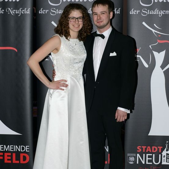 Ein Paar steht lächelnd eng beieinander vor einem Hintergrund mit 'Stadt Neufeld' und einer stilisierten Figur einer Frau. Die Frau trägt ein weißes Kleid und eine Brille, der Mann ist in einem schwarzen Anzug mit Fliege.