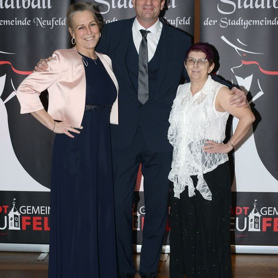 Drei Personen posieren für ein Foto vor einem Hintergrund mit 'Stadt' und 'Gemeinde Neufeld' darauf geschrieben.