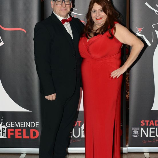 Ein Mann und eine Frau stehen bei einer formellen Veranstaltung eng beieinander und lächeln für ein Foto. Der Mann trägt einen schwarzen Anzug mit einer roten Fliege, während die Frau ein rotes Kleid trägt. Hinter ihnen befinden sich Banner mit Logos und Text.