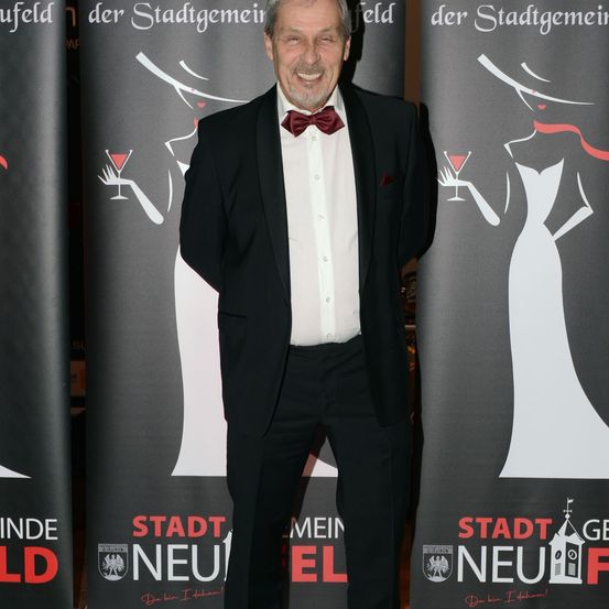 Ein älterer Mann in einem Smoking mit einer roten Fliege steht lächelnd zwischen zwei Bannern.