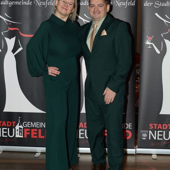 Ein Mann und eine Frau stehen lächelnd nebeneinander bei einer Veranstaltung. Sie sind in passenden grünen Anzügen gekleidet. Hinter ihnen befinden sich Banner mit Text und Bildern.