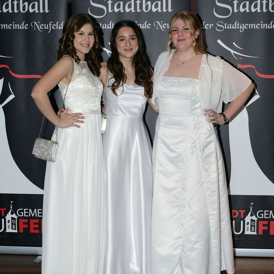 Drei Frauen in weißen Kleidern stehen eng zusammen auf einer Bühne, wahrscheinlich bei einer besonderen Veranstaltung. Die Frau links hält eine silberne Handtasche, und die Frau in der Mitte trägt eine Halskette. Hinter ihnen befindet sich ein schwarzer Hintergrund mit rotem Text und Logos.