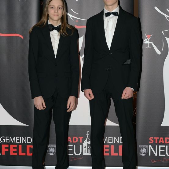 Zwei junge Männer stehen vor einem Hintergrund mit Logos und Text, beide in formellen schwarzen Anzügen mit Fliegen. Der Hintergrund zeigt die Silhouette einer Frau.