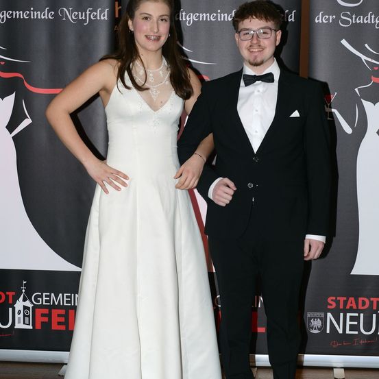 Ein Mann und eine Frau in formeller Kleidung stehen dicht beieinander auf einer Bühne. Die Frau trägt ein weißes Kleid und der Mann einen schwarzen Anzug mit Fliege. Sie stehen vor einem schwarzen Hintergrund mit rotem und weißem Text.