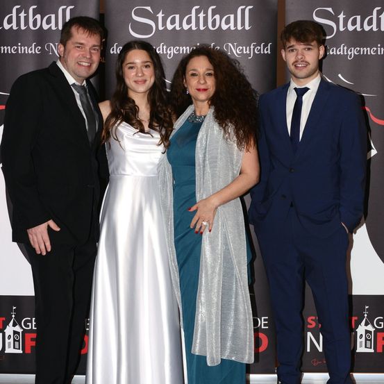 Vier Menschen stehen lächelnd bei einer Veranstaltung zusammen. Ein Mann und eine Frau in formeller Kleidung auf der linken Seite und eine Frau mit einem silbernen Schal und einem Mann in einem marineblauen Anzug auf der rechten Seite. Sie stehen vor einem Hintergrund mit dem Wort 'Stadtball'.