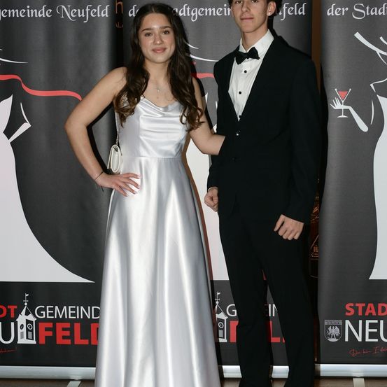 Ein junges Paar in Abendkleidung steht auf einem roten Teppich und lächelt in die Kamera. Die Frau trägt ein silbernes Kleid und der Mann einen schwarzen Anzug mit Fliege. Sie stehen vor einer Kulisse mit rotem Text und einer Silhouette einer Frau.