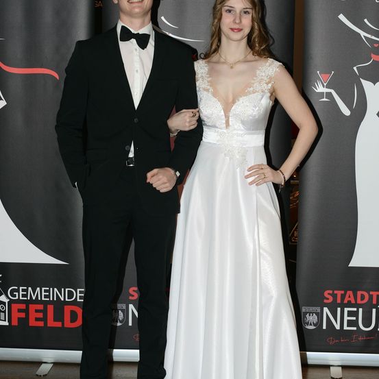 Ein Mann und eine Frau in formeller Kleidung stehen gemeinsam für ein Foto bei einer Veranstaltung. Der Mann trägt einen schwarzen Anzug mit Fliege, während die Frau ein weißes Kleid trägt. Sie stehen vor einem schwarzen Hintergrund mit rotem Text.