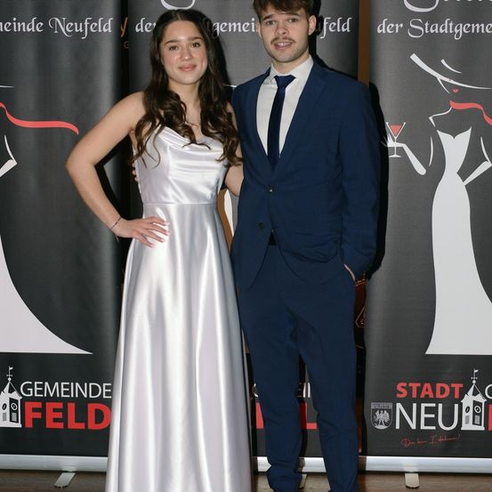 Ein junges Paar posiert in formeller Kleidung vor einem Hintergrund mit rotem und weißem Text.