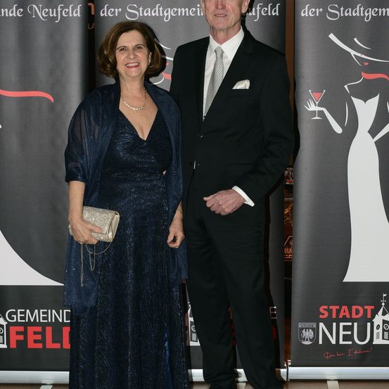 Ein Mann und eine Frau in formeller Kleidung stehen bei einer Veranstaltung nebeneinander. Die Frau hält eine silberne Clutch-Tasche, und der Mann hat ein weißes Einstecktuch. Sie lächeln und blicken in die Kamera. Hinter ihnen befindet sich ein Banner mit dem Text 'STADT NEURFEL'.