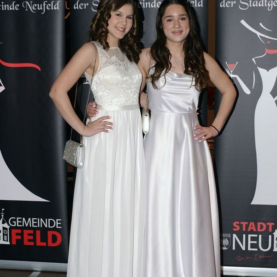 Zwei Frauen stehen eng beieinander und tragen beide weiße Kleider. Sie lächeln und posieren für ein Foto. Hinter ihnen befinden sich zwei Banner. Die Frau auf der linken Seite hält eine Handtasche.